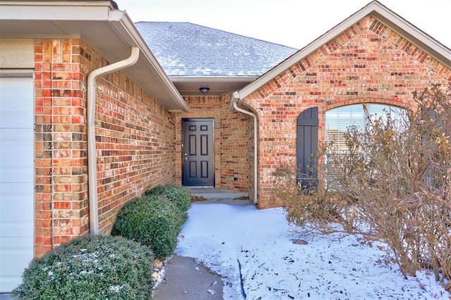 snow covered property entrance featuring a garage, brick siding, and a shingled roof