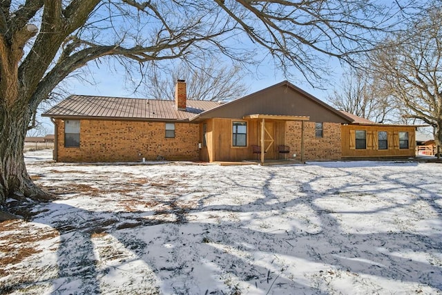 snow covered rear of property with brick siding, metal roof, and a chimney