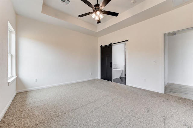 unfurnished bedroom featuring carpet, ensuite bath, a barn door, ceiling fan, and a raised ceiling