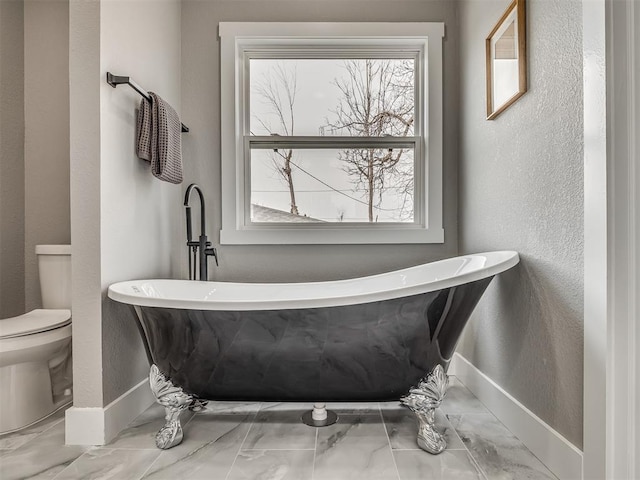 bathroom featuring a textured wall, baseboards, a freestanding bath, and toilet