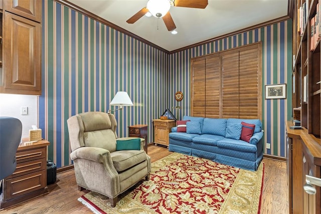 living area featuring wallpapered walls, baseboards, ceiling fan, ornamental molding, and wood finished floors