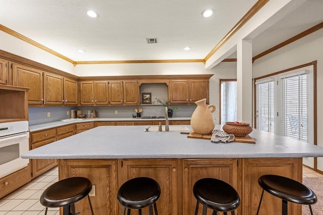 kitchen with brown cabinetry, oven, a kitchen island with sink, light countertops, and a kitchen bar