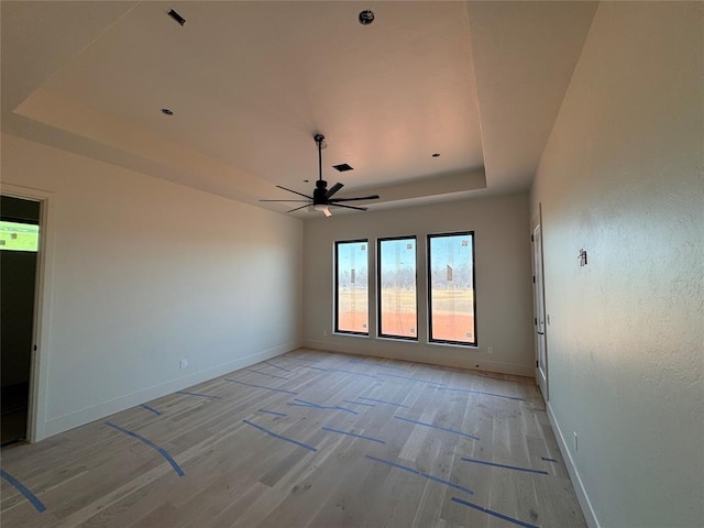 empty room with light wood-style floors, a raised ceiling, baseboards, and a ceiling fan