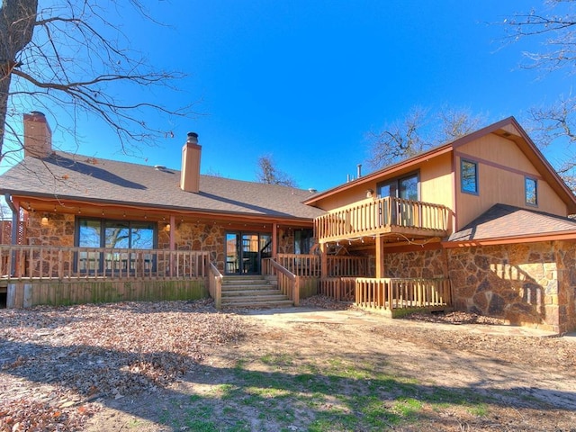 rear view of house featuring stone siding, roof with shingles, a chimney, and a wooden deck