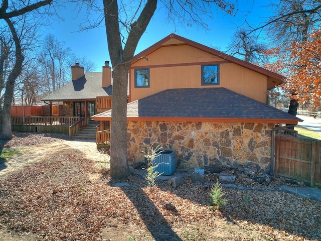 back of house with central AC unit, a shingled roof, fence, stone siding, and a chimney