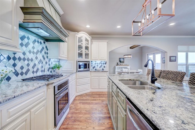 kitchen with arched walkways, stainless steel appliances, custom range hood, light wood-style floors, and a sink