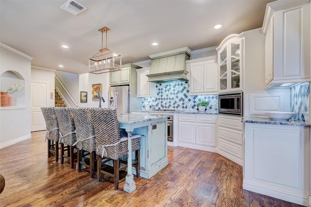 kitchen with custom exhaust hood, stainless steel appliances, a kitchen island, light stone countertops, and a kitchen breakfast bar