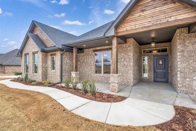 property entrance featuring brick siding, covered porch, and roof with shingles