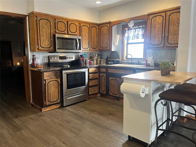 kitchen featuring brown cabinetry, dark wood finished floors, dark countertops, stainless steel appliances, and a sink