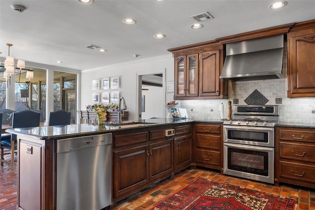 kitchen with wall chimney range hood, decorative backsplash, appliances with stainless steel finishes, a peninsula, and a sink