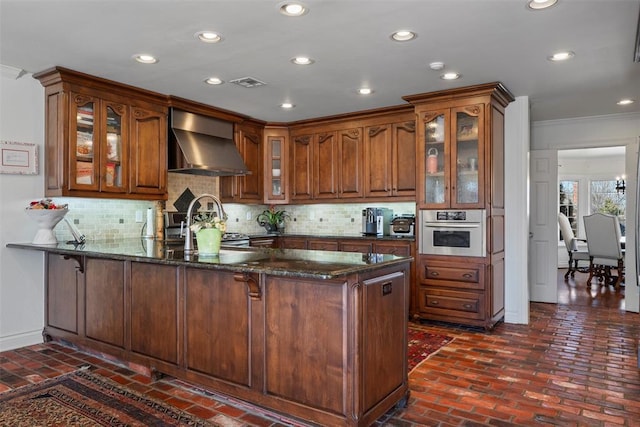 kitchen featuring visible vents, a peninsula, brick floor, stainless steel oven, and wall chimney range hood