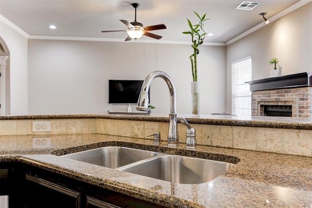 kitchen featuring crown molding, visible vents, a brick fireplace, a sink, and ceiling fan