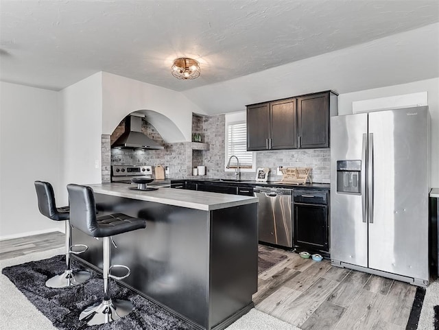 kitchen featuring a breakfast bar, stainless steel appliances, wall chimney exhaust hood, and light wood-style floors