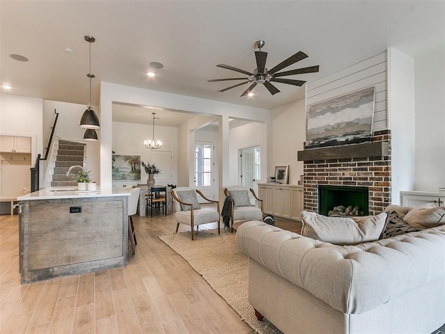 living room featuring ceiling fan with notable chandelier, stairway, a brick fireplace, and light wood-style flooring