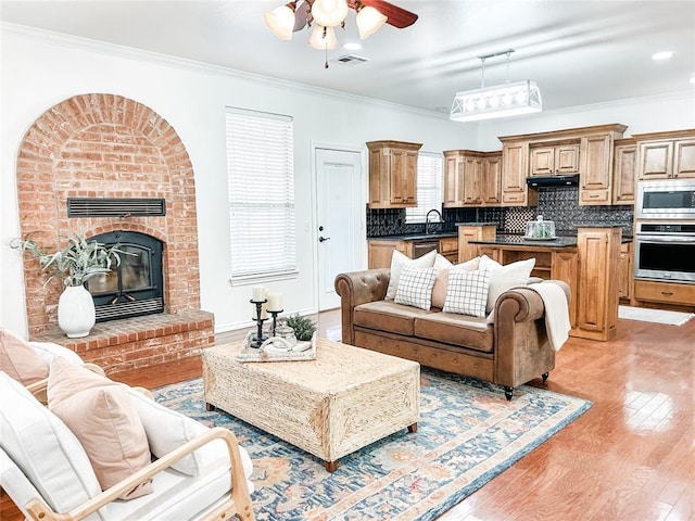 living room with a brick fireplace, visible vents, a ceiling fan, ornamental molding, and light wood-style floors