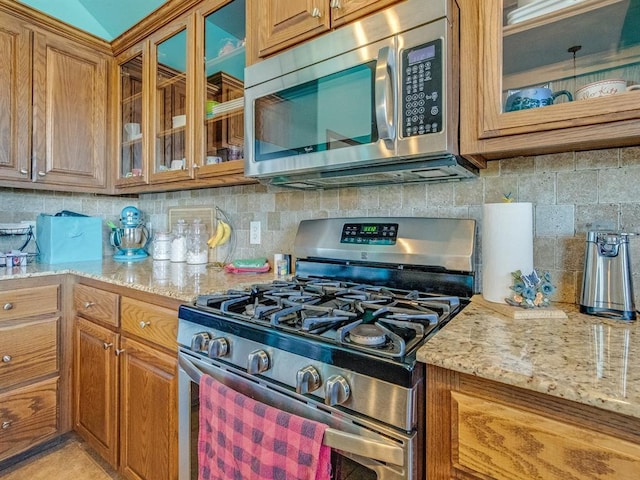 kitchen with tasteful backsplash, brown cabinetry, glass insert cabinets, light stone countertops, and stainless steel appliances