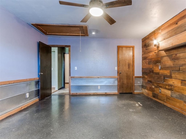 unfurnished room featuring attic access, wood walls, a ceiling fan, and speckled floor
