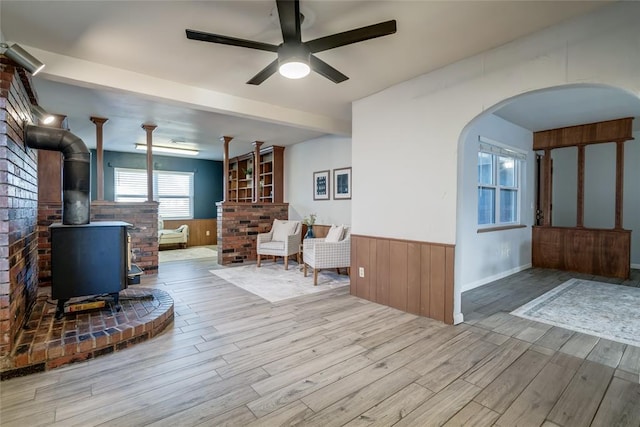 unfurnished living room with arched walkways, a wainscoted wall, a wood stove, ceiling fan, and wood finished floors