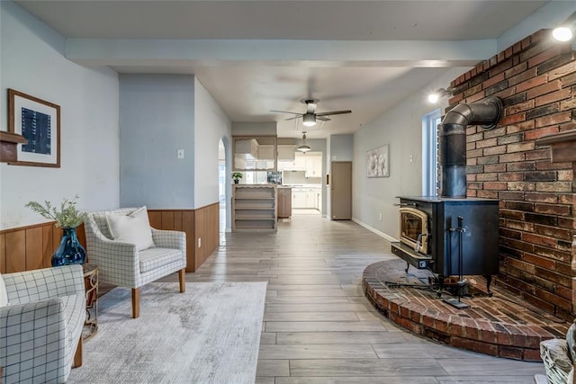 living area with a wainscoted wall, ceiling fan, wood finished floors, beamed ceiling, and a wood stove