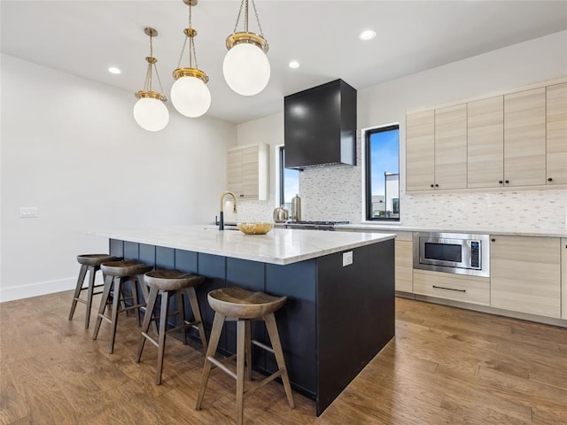 kitchen featuring a kitchen island with sink, a breakfast bar, wood finished floors, wall chimney range hood, and stainless steel microwave