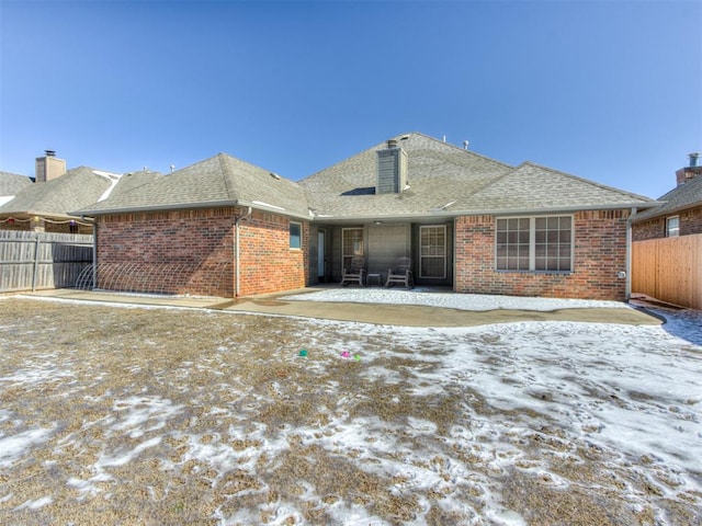 snow covered rear of property featuring a patio area, a shingled roof, fence, and brick siding