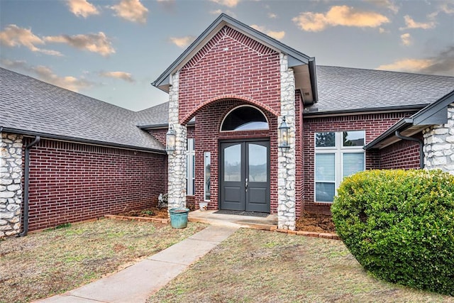 view of exterior entry with stone siding, brick siding, french doors, and roof with shingles