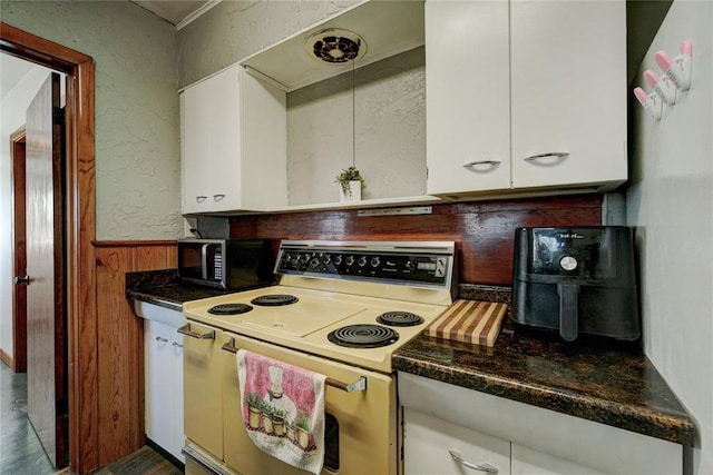 kitchen with a textured wall, wood walls, dark stone counters, white cabinets, and electric range oven