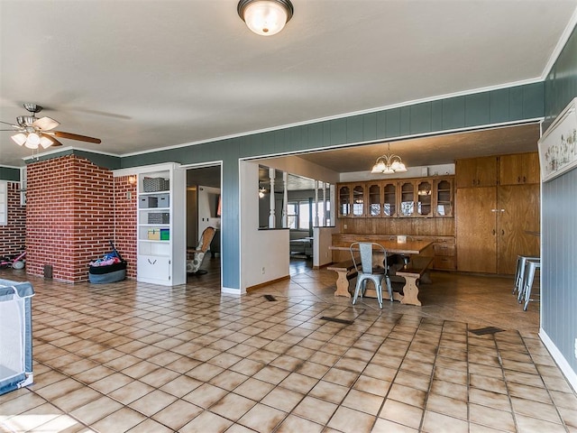 unfurnished dining area with baseboards, brick wall, ceiling fan with notable chandelier, crown molding, and light tile patterned flooring