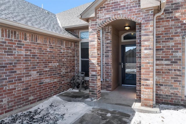 snow covered property entrance featuring a shingled roof and brick siding