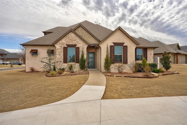 french country style house featuring stucco siding, roof with shingles, a front yard, and brick siding