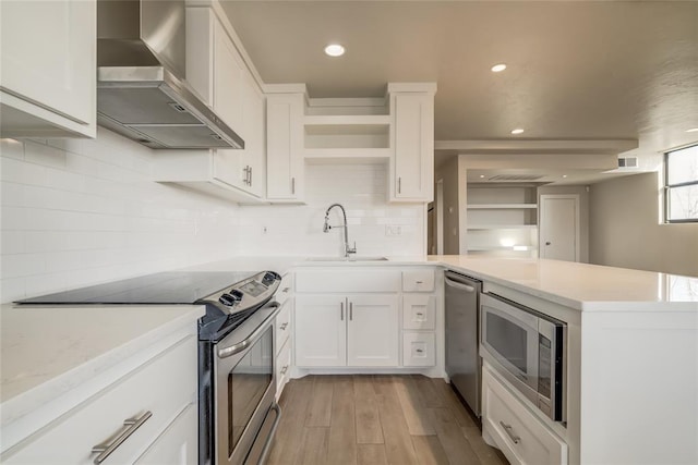 kitchen with light countertops, appliances with stainless steel finishes, white cabinets, a sink, and wall chimney range hood