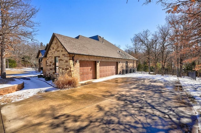 view of snowy exterior featuring a shingled roof, central AC unit, concrete driveway, stone siding, and fence