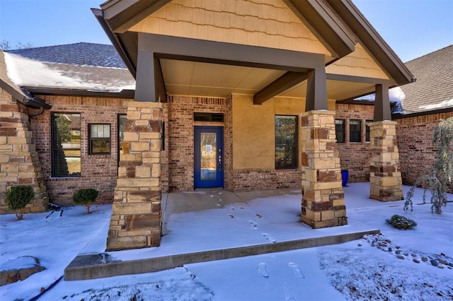 snow covered property entrance with stone siding, brick siding, and roof with shingles