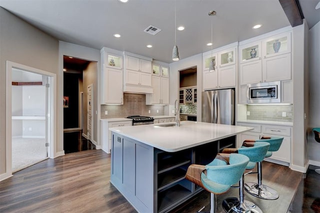 kitchen featuring stainless steel appliances, white cabinets, light countertops, an island with sink, and glass insert cabinets