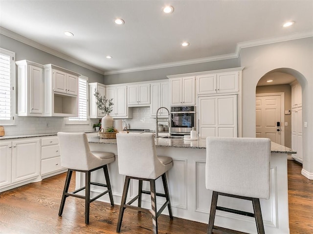 kitchen featuring dark wood-type flooring, a center island with sink, stainless steel oven, arched walkways, and white cabinetry