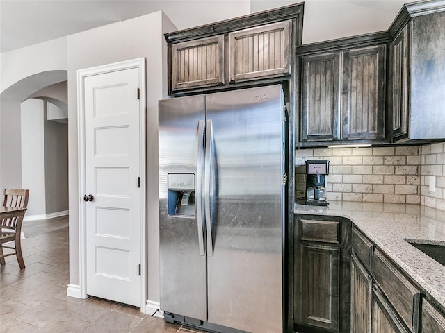 kitchen featuring arched walkways, dark brown cabinetry, stainless steel fridge with ice dispenser, and light stone countertops