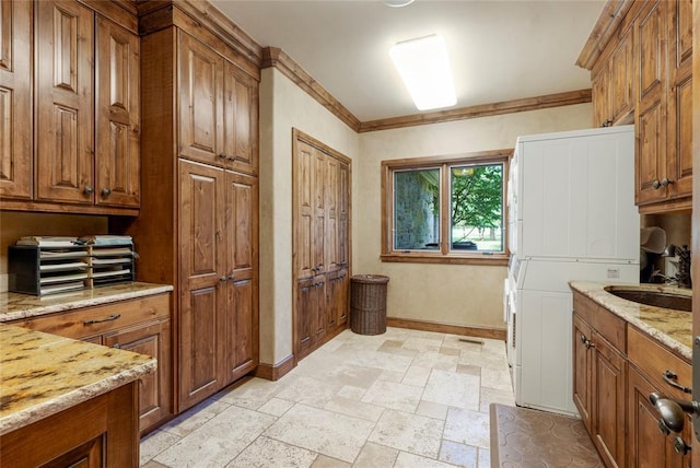 kitchen featuring brown cabinetry, stacked washer / drying machine, light stone countertops, stone tile flooring, and a sink