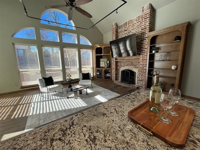 living room featuring baseboards, ceiling fan, wood finished floors, a brick fireplace, and high vaulted ceiling