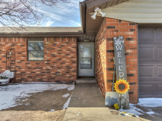 snow covered property entrance with a garage, brick siding, and roof with shingles