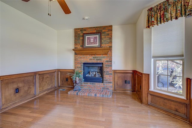 living area featuring a ceiling fan, a brick fireplace, a wainscoted wall, and wood finished floors