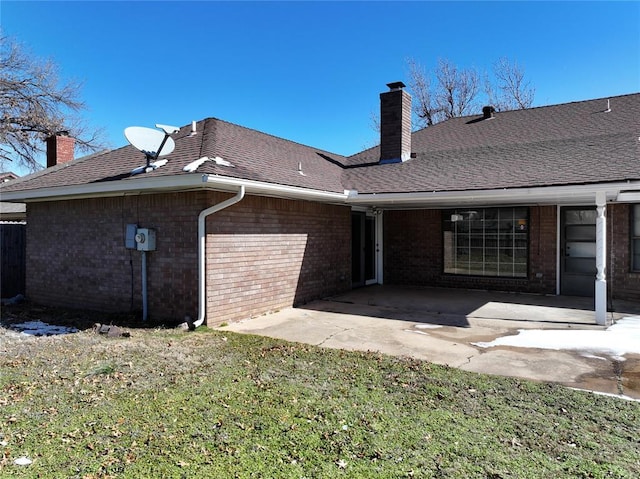 back of property with brick siding, roof with shingles, a chimney, a lawn, and a patio area