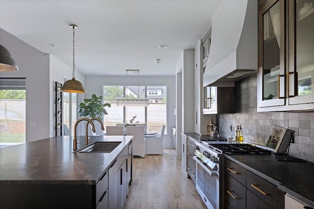 kitchen with custom exhaust hood, stainless steel stove, glass insert cabinets, a kitchen island with sink, and a sink