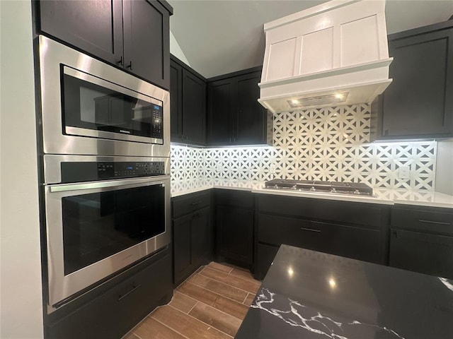 kitchen with dark cabinetry, wood tiled floor, custom exhaust hood, stainless steel appliances, and decorative backsplash