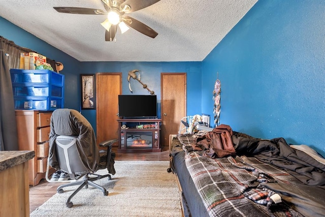 bedroom featuring light wood-style flooring, a ceiling fan, a textured ceiling, and a glass covered fireplace