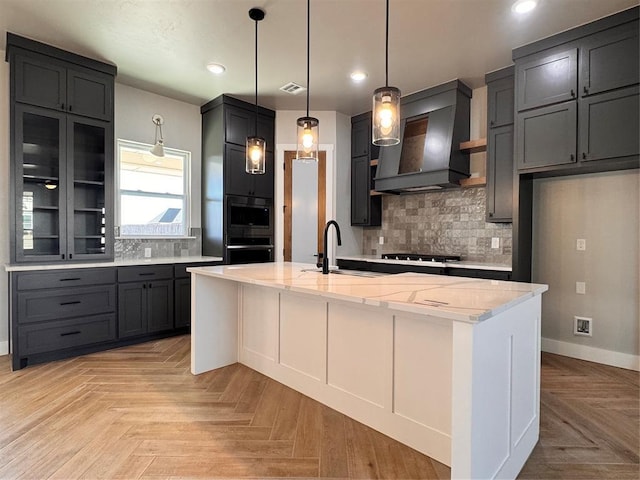 kitchen with a sink, a kitchen island with sink, custom exhaust hood, and light stone counters