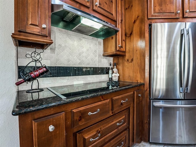 kitchen featuring black electric cooktop, under cabinet range hood, freestanding refrigerator, decorative backsplash, and dark stone countertops