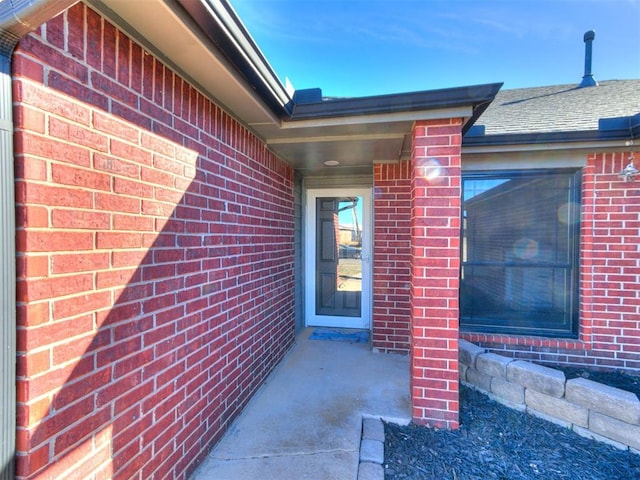 view of exterior entry with roof with shingles and brick siding