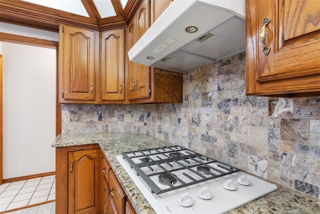 kitchen with light tile patterned flooring, under cabinet range hood, white gas stovetop, tasteful backsplash, and brown cabinetry