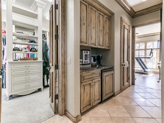 kitchen featuring light tile patterned floors, light colored carpet, brown cabinetry, dark stone countertops, and crown molding