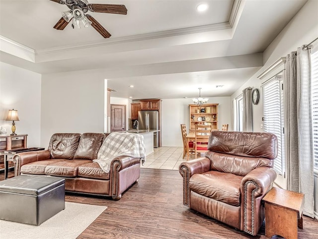 living area featuring a raised ceiling, crown molding, light wood-style flooring, and ceiling fan with notable chandelier